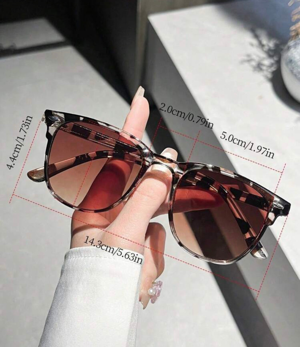 Sunglasses held by a hand with measurements on a gray surface