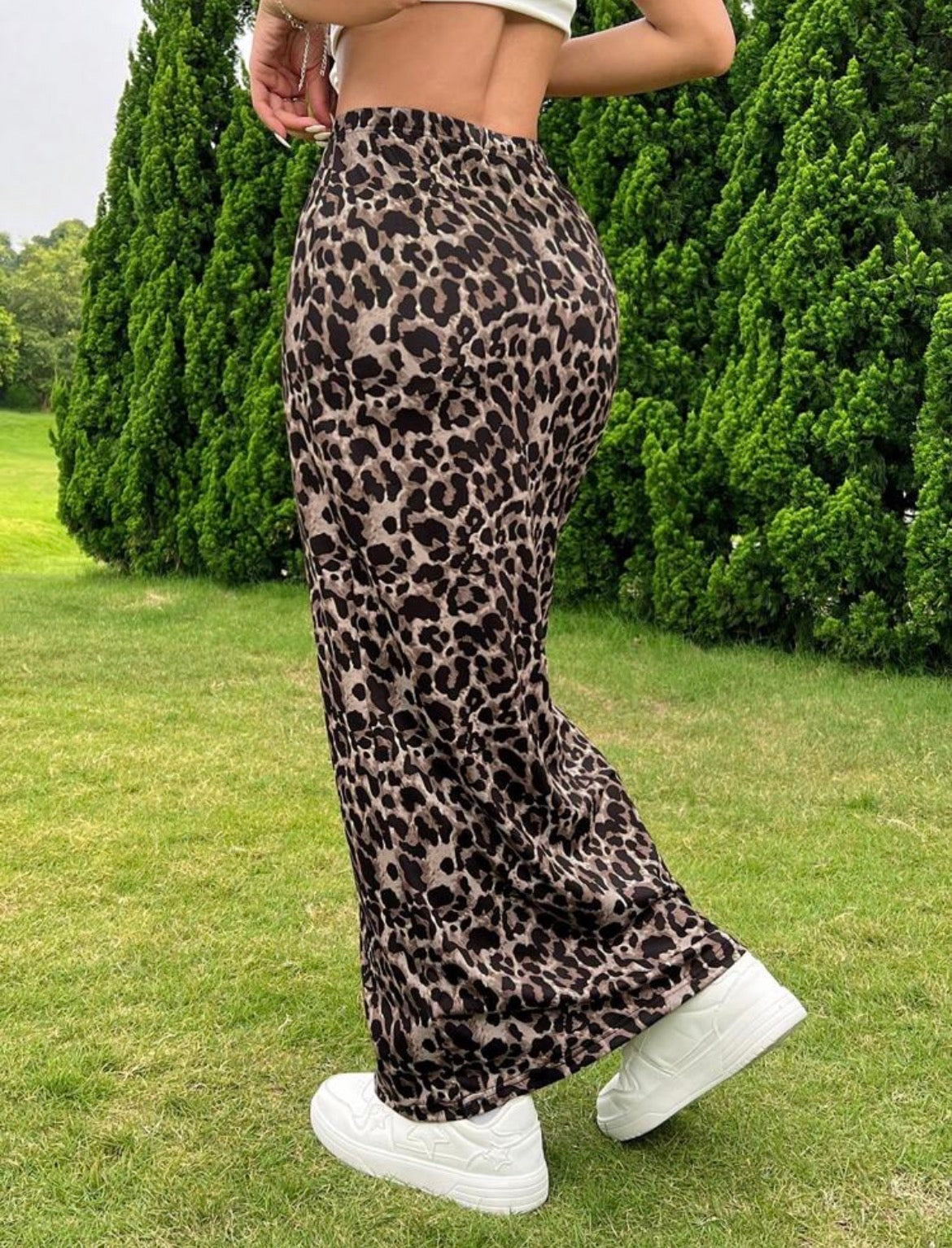 Person wearing leopard print pants and white sneakers in a grassy area with green bushes.