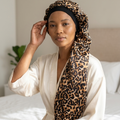 Woman wearing a leopard print sleep cap and white robe on a bed.